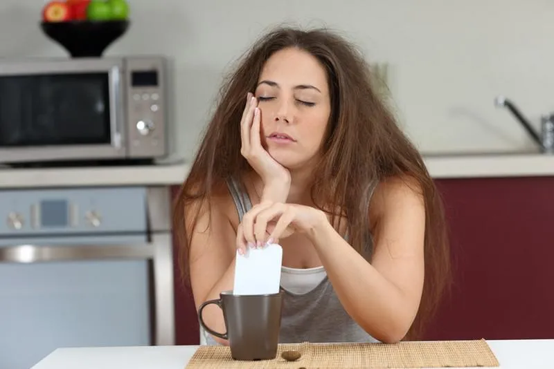 fille fatiguée endormie avec un mauvais réveil en remuant le café avec le téléphone assis dans la cuisine à la maison