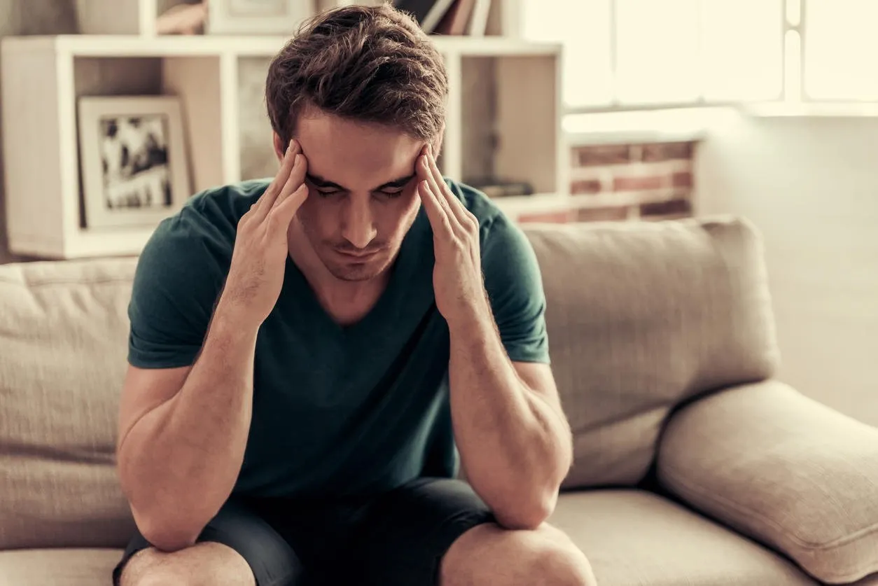 handsome tired guy in casual clothes is massaging his temples while sitting on couch at home