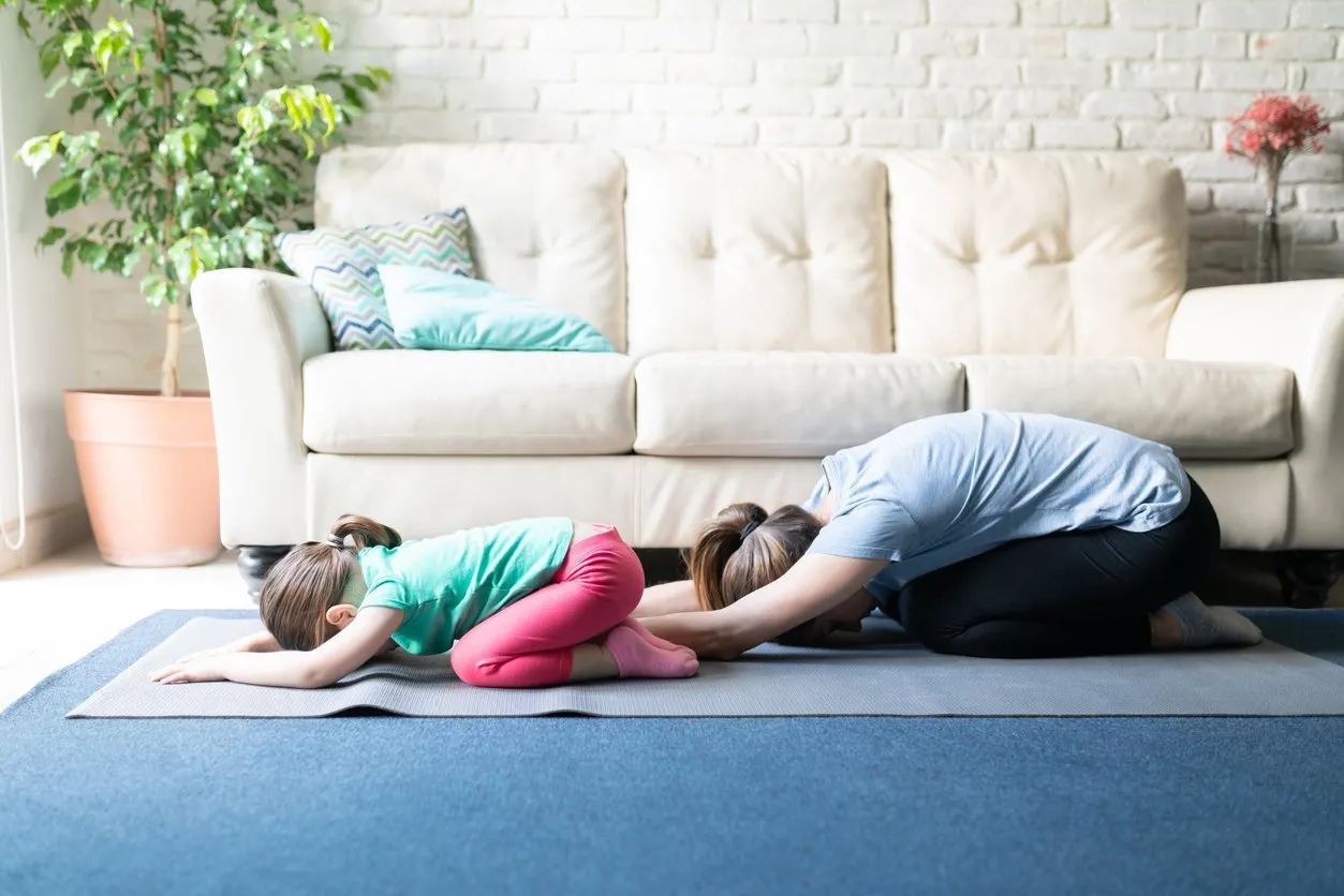 both mother and daughter exercising together and doing yoga child's pose in the living room at home