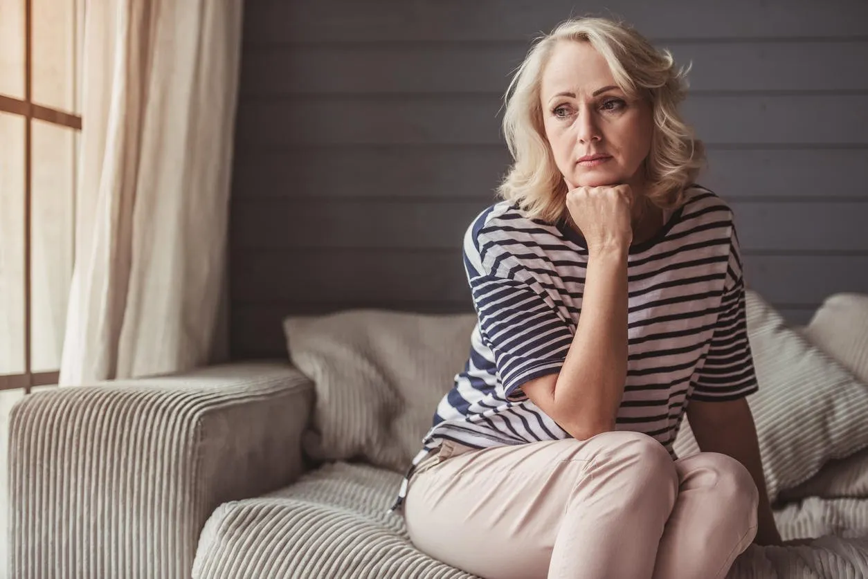 beautiful sad senior woman is leaning on her hand and looking downward while sitting on couch at home