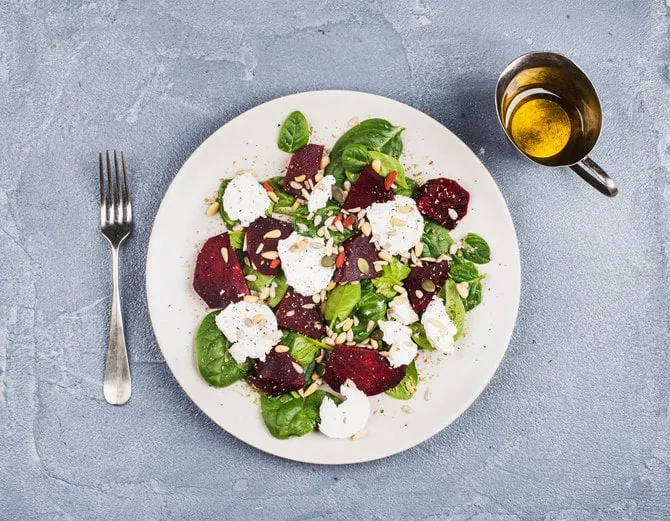 salad with roasted beetroot, spinach, soft goat heese and seeds in light plate over grey concrete textured background, oil in saucer top view