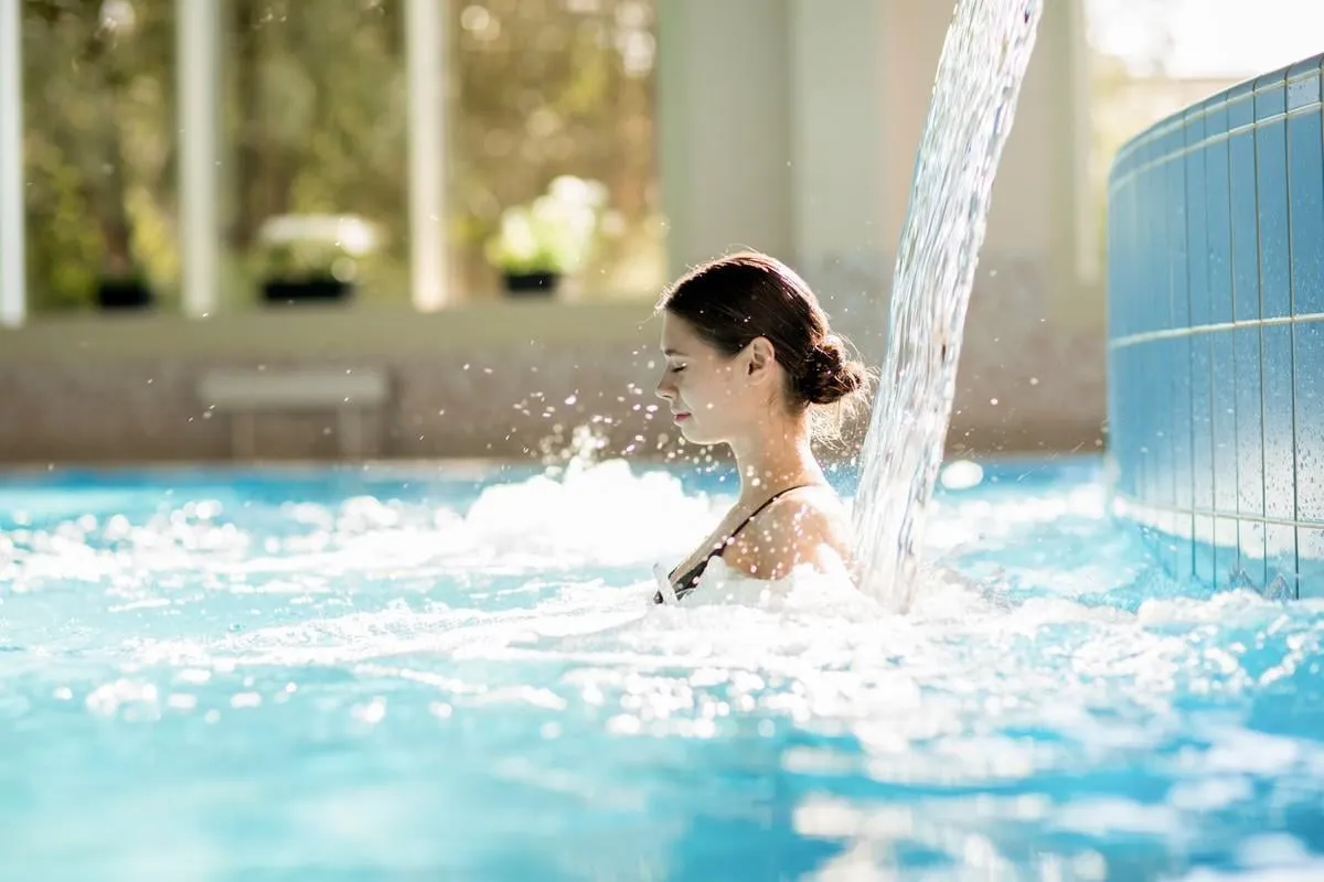 serene girl enjoying stream of waterfall and its gentle splashes in swimming-pool at spa resort