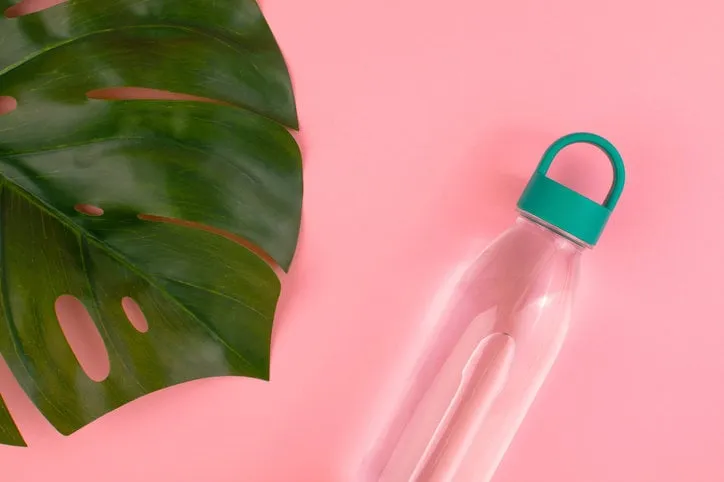 reusable plastic water bottle with green cap and big leaf of tropical plant monstera at pink background top view, copy space