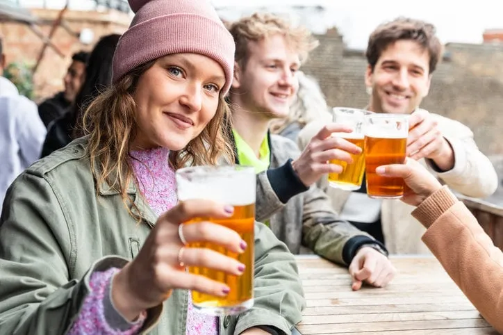 friends drinking and toasting with beer at pub brewery - beautiful young woman looking at camera and raising her pint glass - lifestyle and drink concepts in london