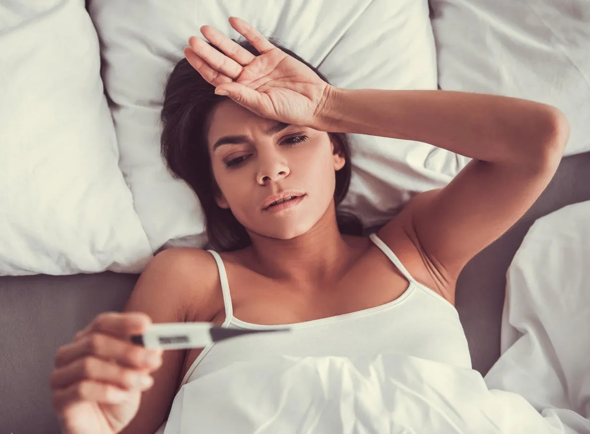young beautiful woman lying in bed, she measures her temperature with a thermometer and touching her forehead