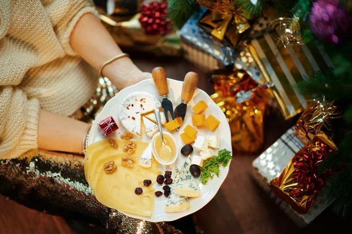 closeup on trendy middle age woman in gold sequin skirt and white sweater under decorated christmas tree near present boxes holding cheese platter
