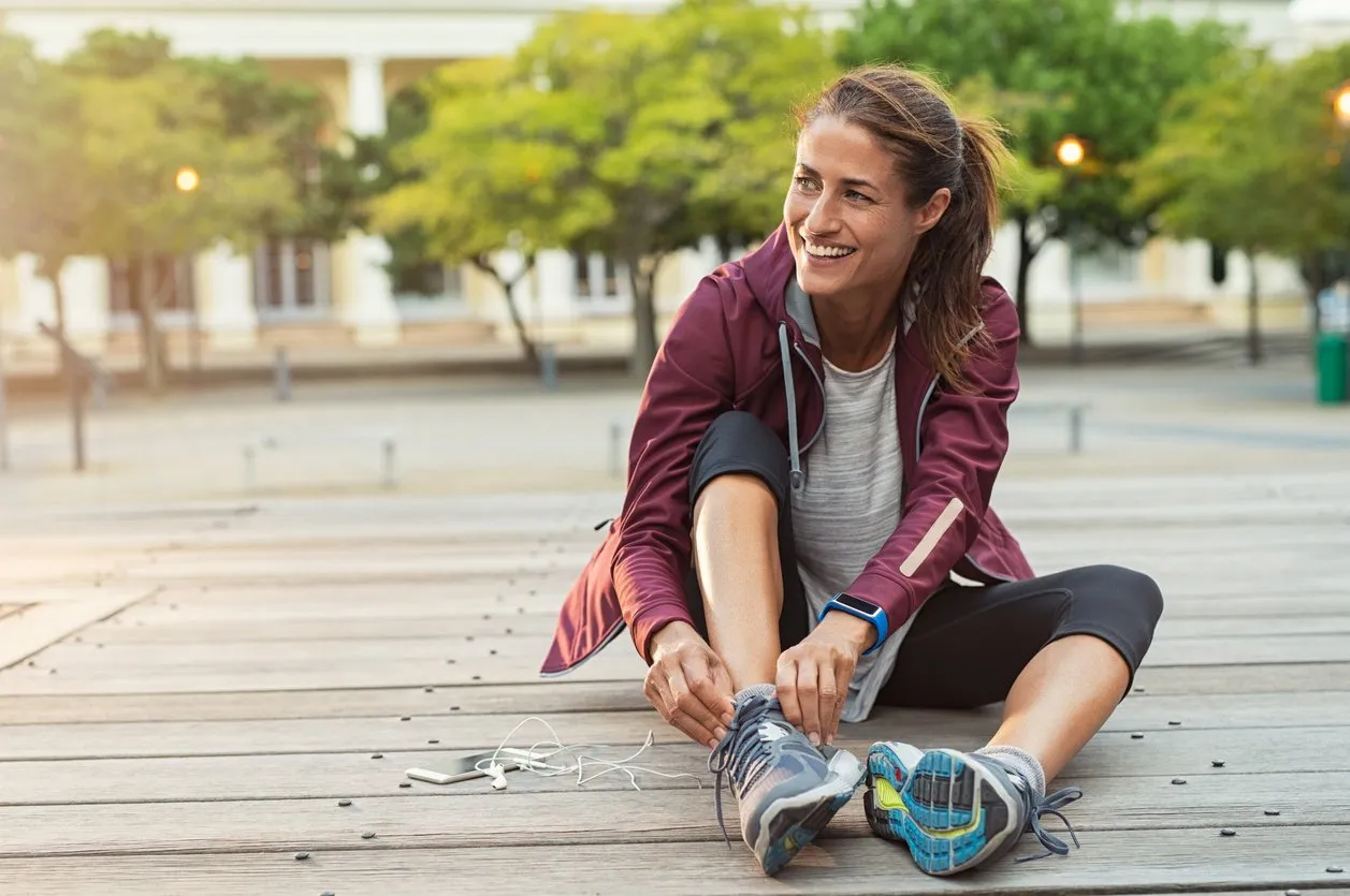 mature fitness woman tie shoelaces on road cheerful runner sitting on floor on city streets with mobile and earphones wearing sport shoes active latin woman tying shoe lace before running