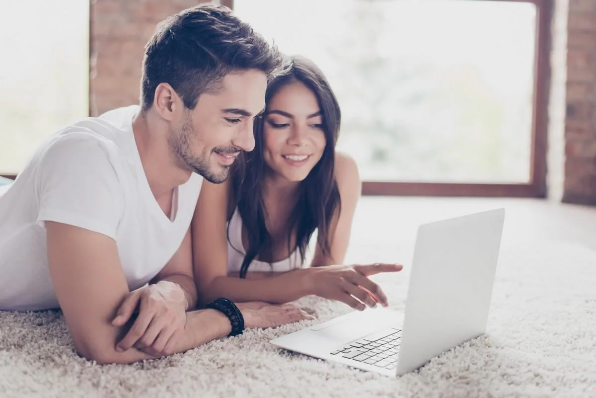 beautiful latino mulatto lovers are browsing info on laptop and discussing it, lying on the floor on comfortable carpet at home they are smiling, so relaxed