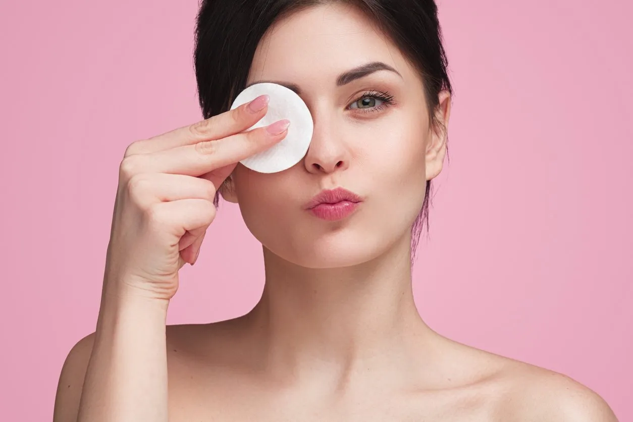 pretty young female wiping eye with cotton pad in studio with pink background and looking at camera