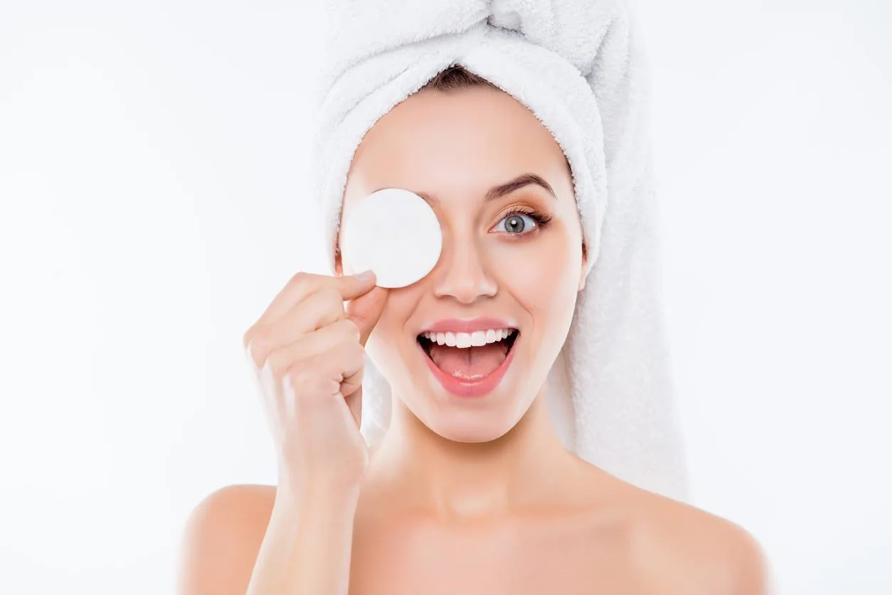 portrait of foolish playful woman using sponge for application of lotion close one eye with cotton keeping open mouth with towel on head isolated on white background