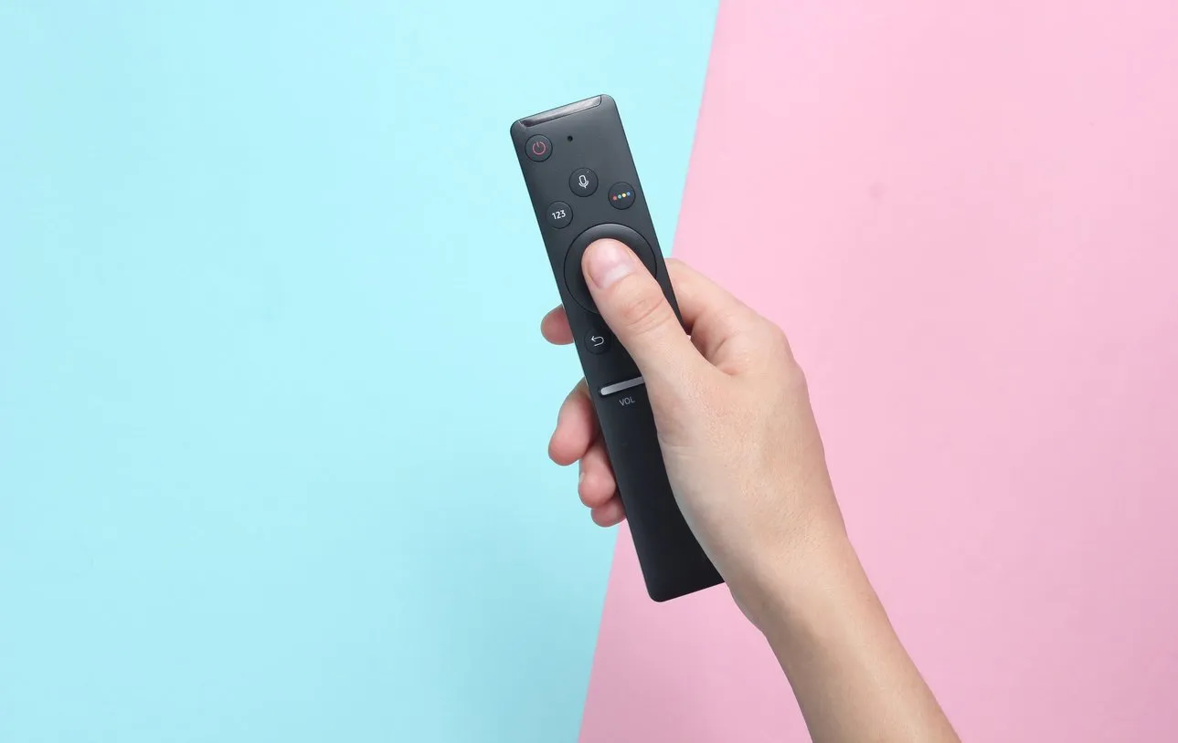female hand holds the tv remote on a pink blue pastel background top view