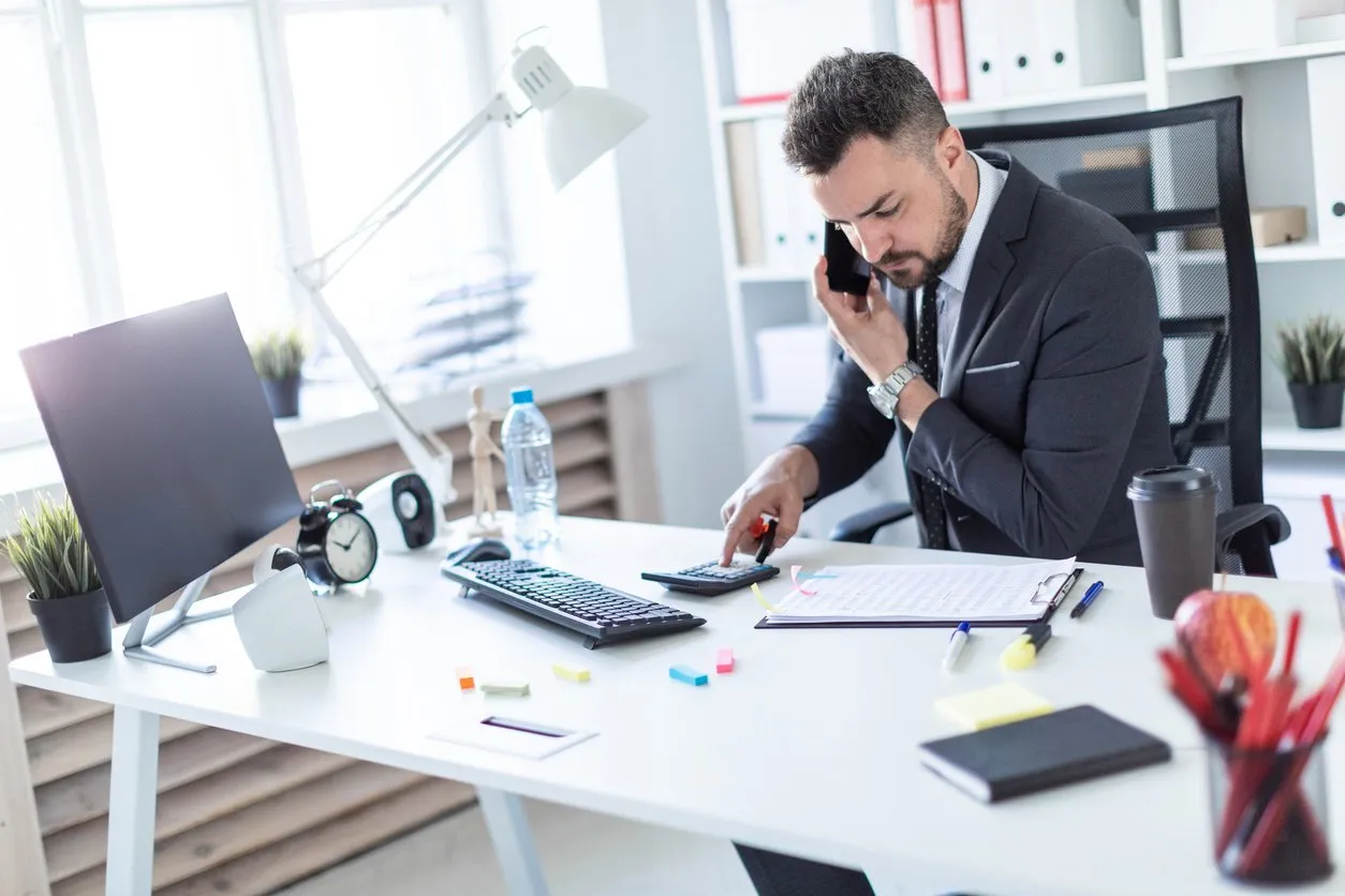 a man is sitting at the desk at the office, talking on the phone, holding a marker in his hand and counting on a calculator
