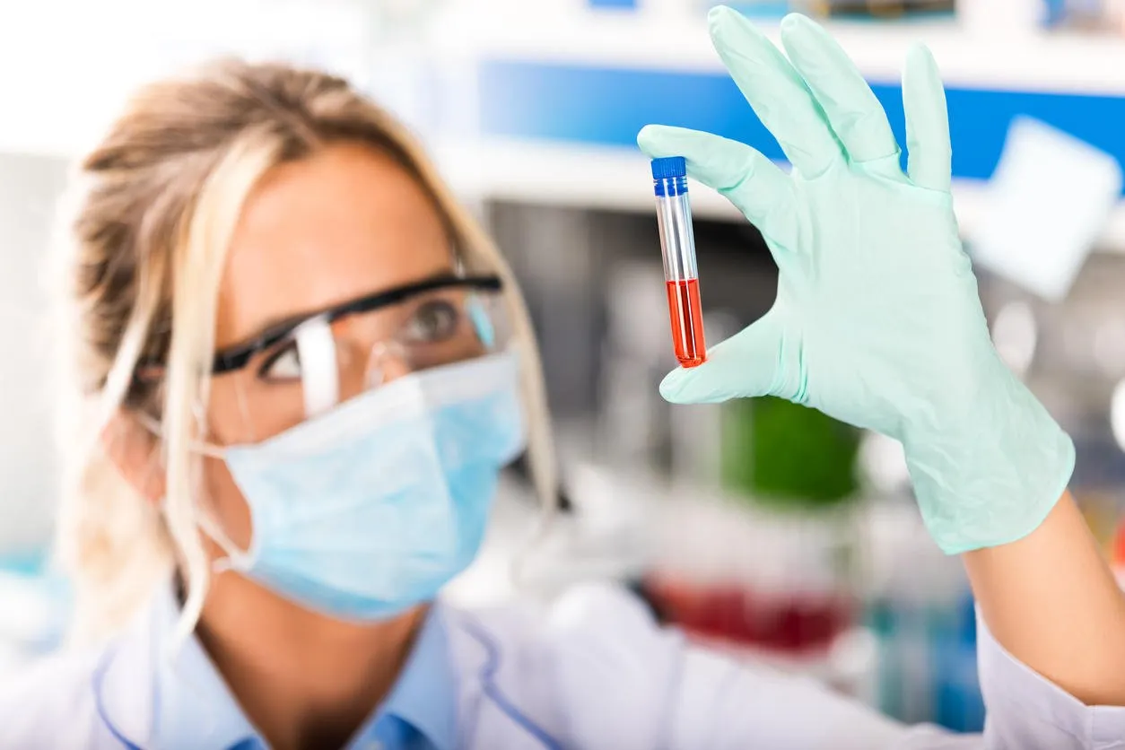 young attractive female scientist in protective eyeglasses and mask examining test tube with red liquid sample substance probe in the scientific chemical research laboratory