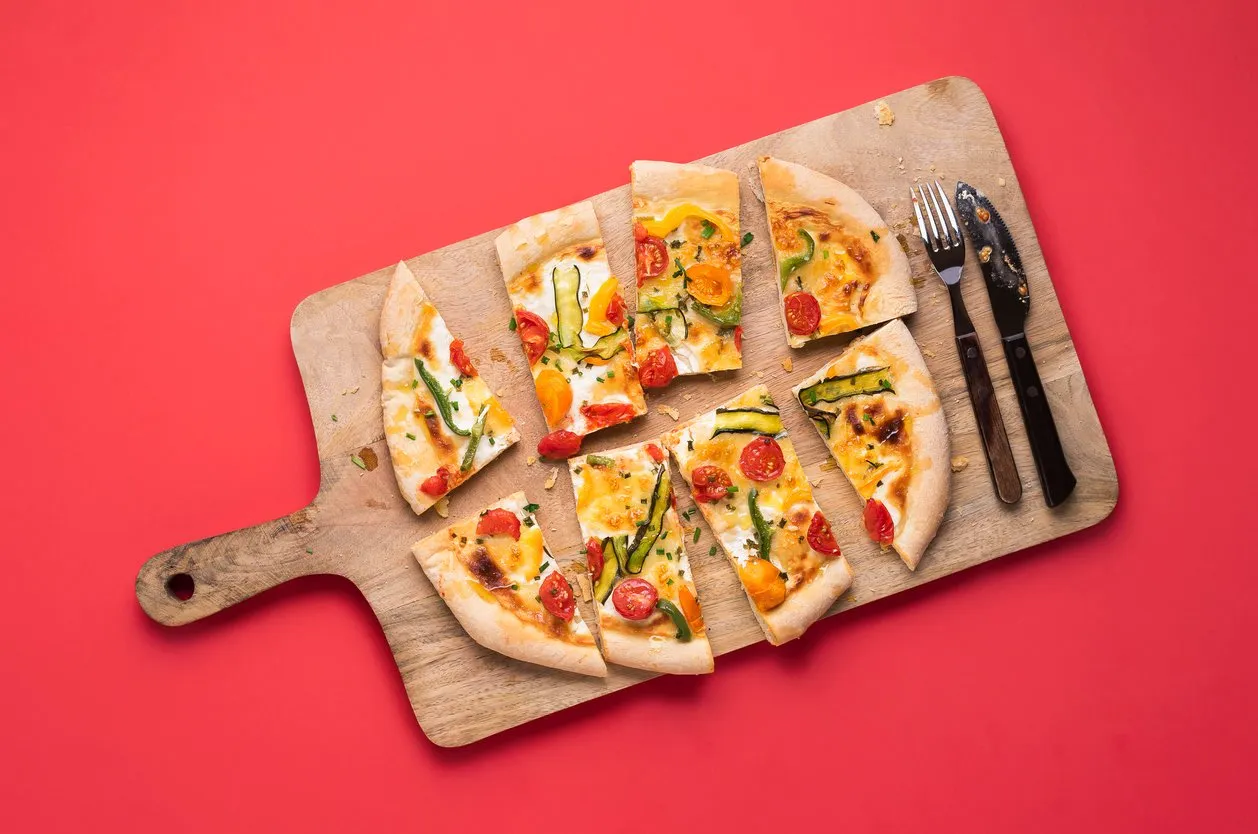 spring pizza with fresh veggies, sliced, on wooden cutting board with cutlery, on red background above view of sliced pizza primavera italian food