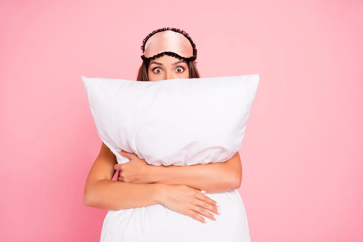 young gorgeous shocked lady wearing eye mask, hugging pillow, hiding isolated over pink pastel background