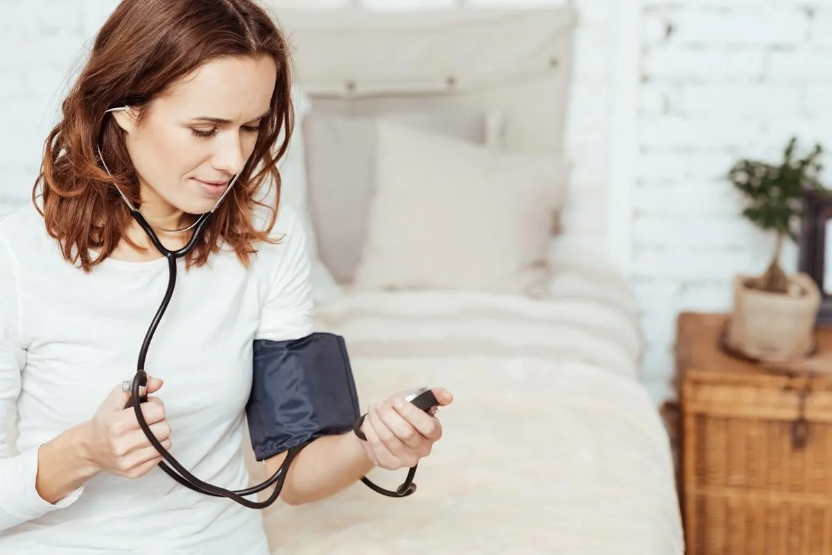 under control pleasant charmign young woamn concentrating on measuring blood pressure and uisng tonometer while sitting on her bed