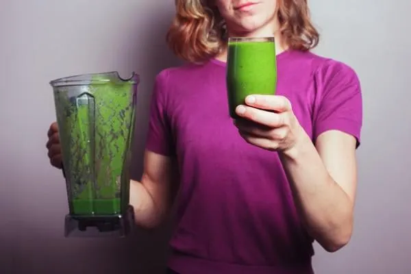 une jeune femme dans un haut violet tient un mélangeur et un verre avec smoothie aux fruits verts