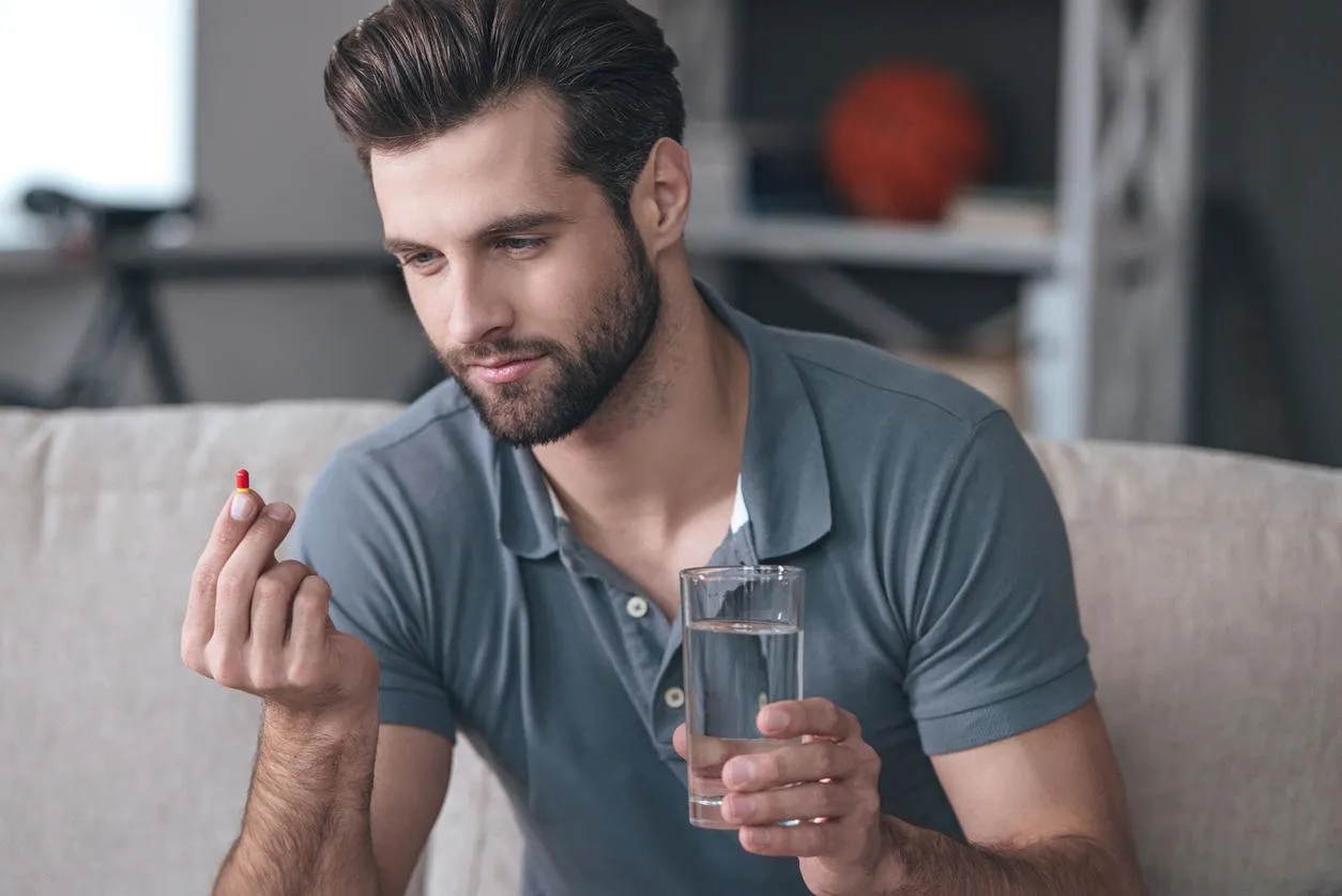 beau jeune homme tenant un verre d'eau et en regardant une pilule dans sa main tout en étant assis sur le canapé à la maison