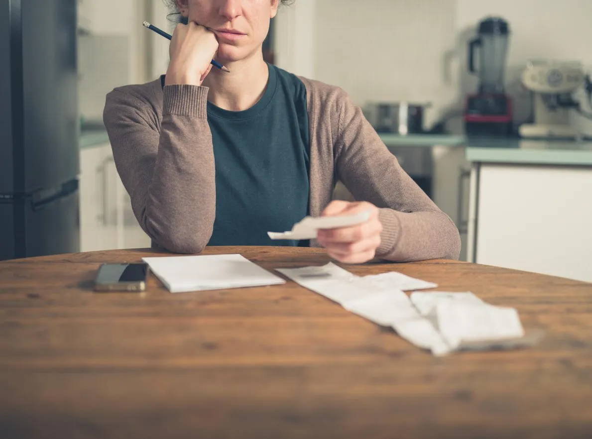 a young woman is sitting in her kitchen and is loking at her receipts at home while using a smart phone