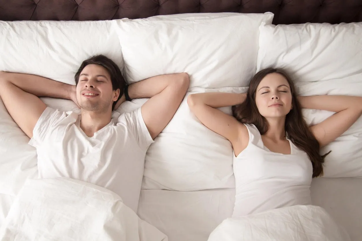 young happy married couple wearing white resting with eyes closed in bed, lying with hands behind the head after waking up in the morning, or going to sleep at night lovers relaxing in bed top view