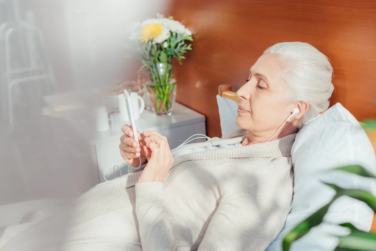 senior woman in earphones using smartphone while lying on hospital bed