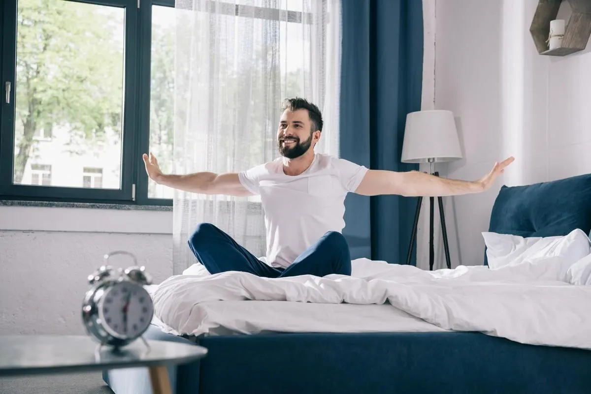 smiling young man in pajamas stretching while sitting on bed at morning