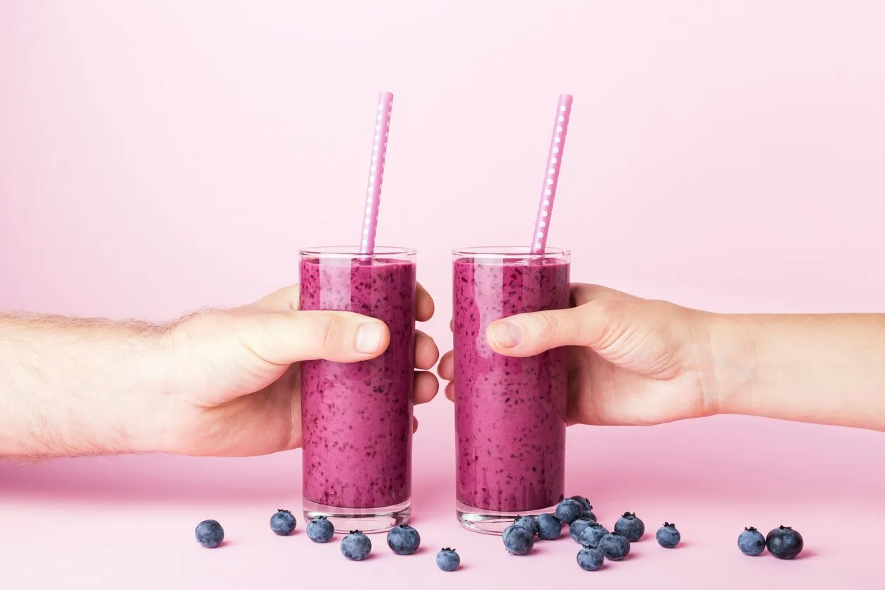 woman's and man's hands holding two glasses of blueberries smoothie with straws on pink background healthy summer drink