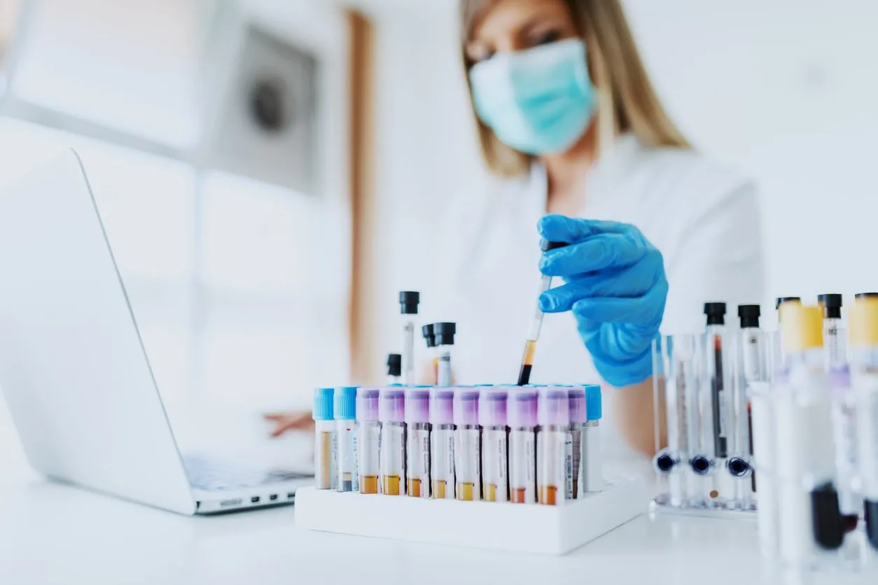close up of lab assistant in uniform, with mask and rubber gloves holding test tube with blood sample while sitting on chair and typing on laptop selective focus on test tubes