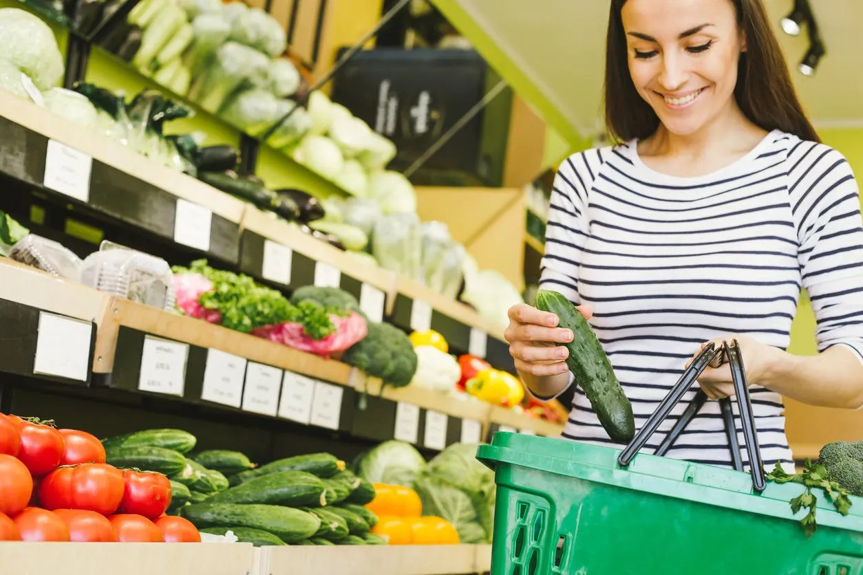 the choice of vegetables in the supermarket smiling beautiful woman puts a cucumber in a food basket on a background of vegetables and fruits
