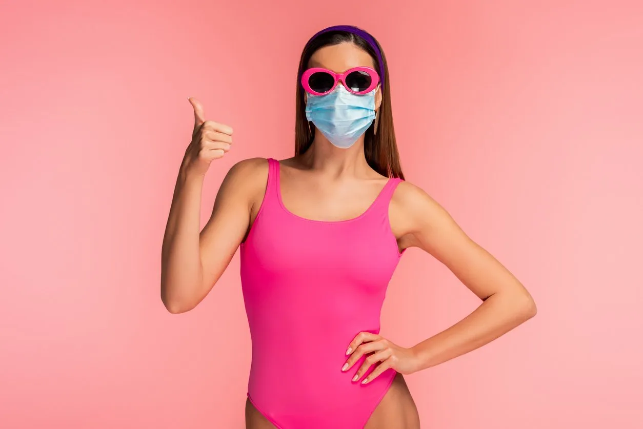 front view of woman with hand on hip and medical mask showing like sign isolated on pink