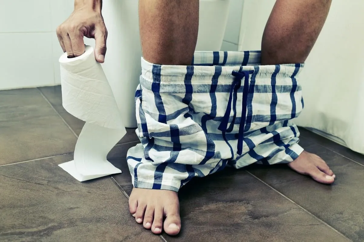 detail of a young caucasian man in the toilet sitting in the bowl, with a roll of toilet paper in his hand