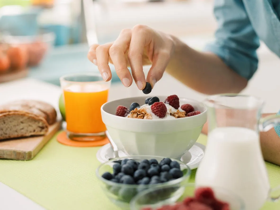 woman having an healthy delicious breakfast at home with yogurt, cereals and fresh fruit, she is picking a blueberry