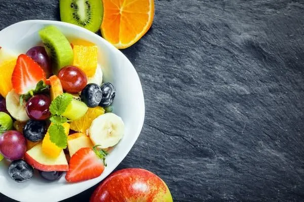 vue aérienne d'un bol de salade de fruits frais pour une alimentation saine avec des fruits tropicaux assortis et une pomme, un kiwi et une orange disposés côte à côte sur une ardoise texturée avec fond