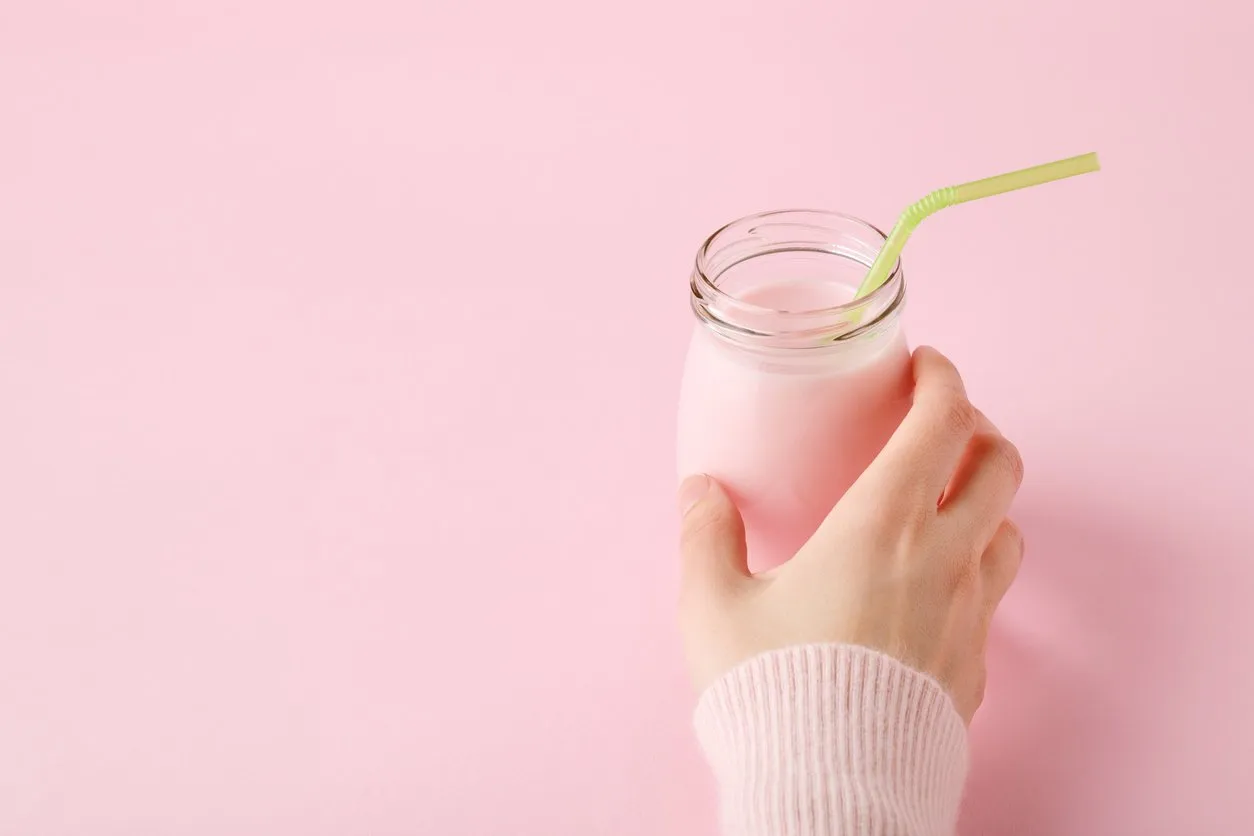 women's hand holding glass bottle of fruit yogurt with straw on pastel pink background with copy space healthy drink concept, minimal