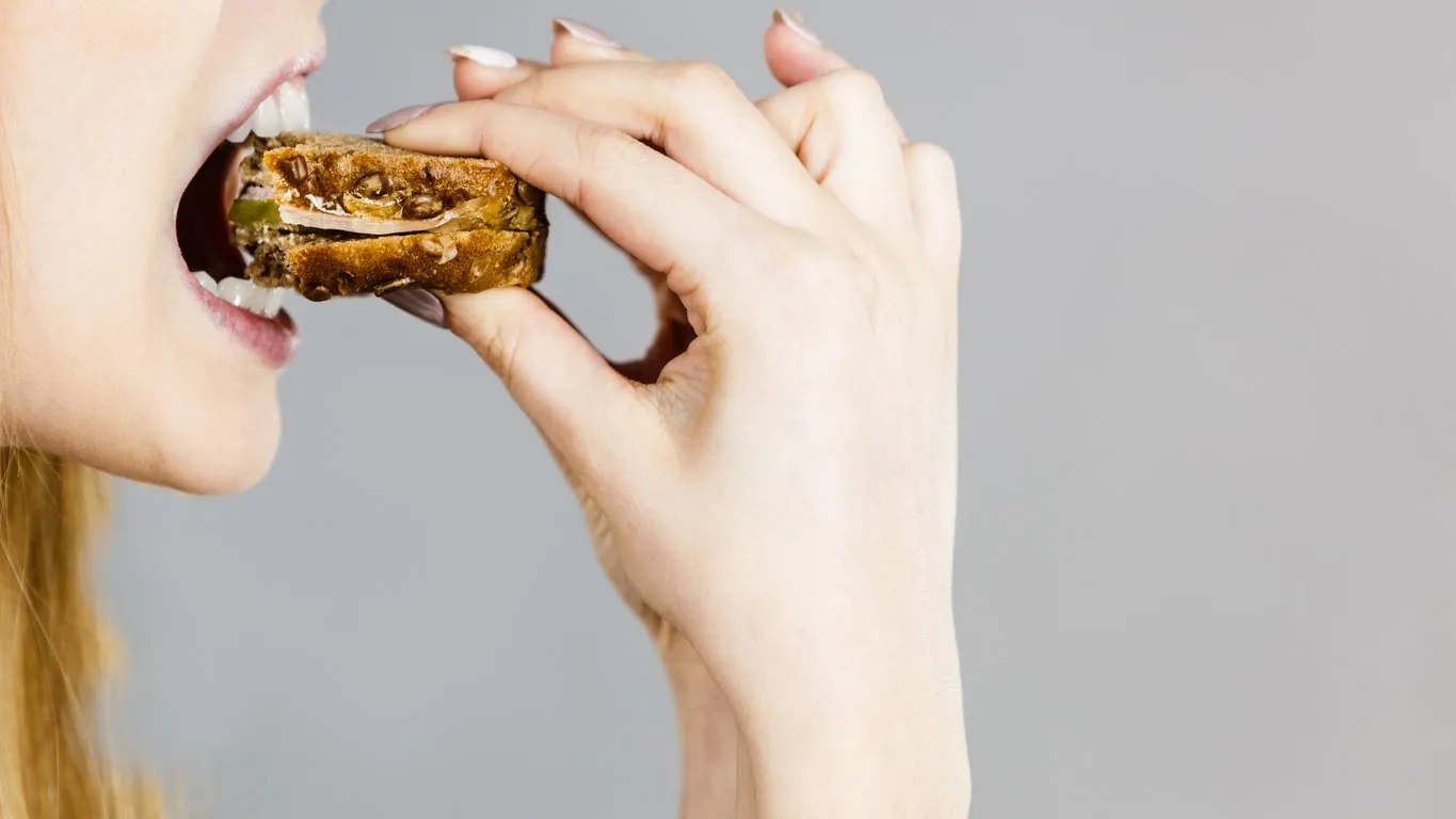 young woman eating sandwich, taking bite with wide open mouth food, calories, dieting concept studio shot on grey background, profile view