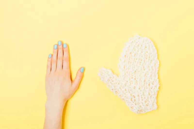 flat lay concept of body care bath accessories on bright yellow background minimal composition of woman's hand with blue manicure next to coarse mitten washcloth