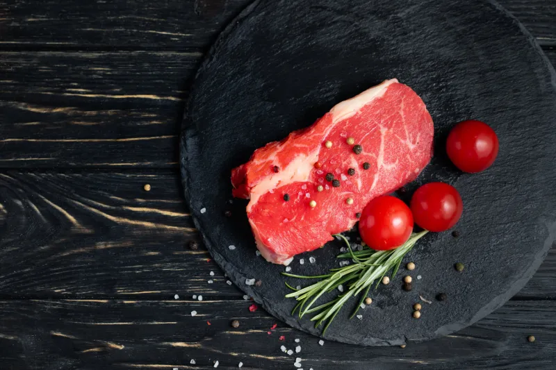 one pieces of juicy raw beef with rosemary on a stone cutting board on a black wooden table background meat for barbecue or grill next to cherry tomatoes sprinkled with pepper and salt seasoning