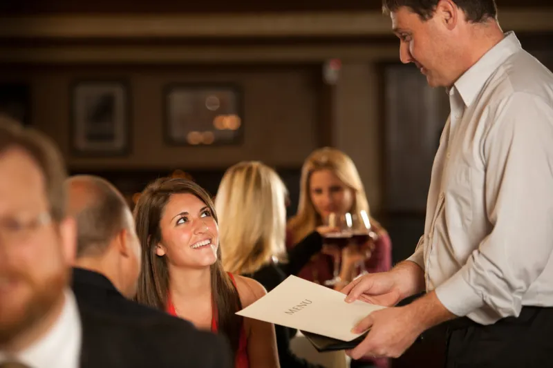 scene in a crowded restaurant as a male waiter brings a menu to a brunette woman she looks up and smiles