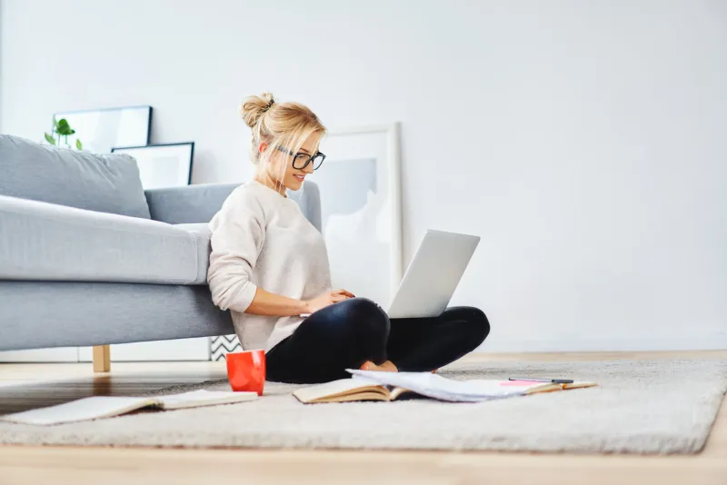 female student sitting on floor of her apartment with laptop and notes studying