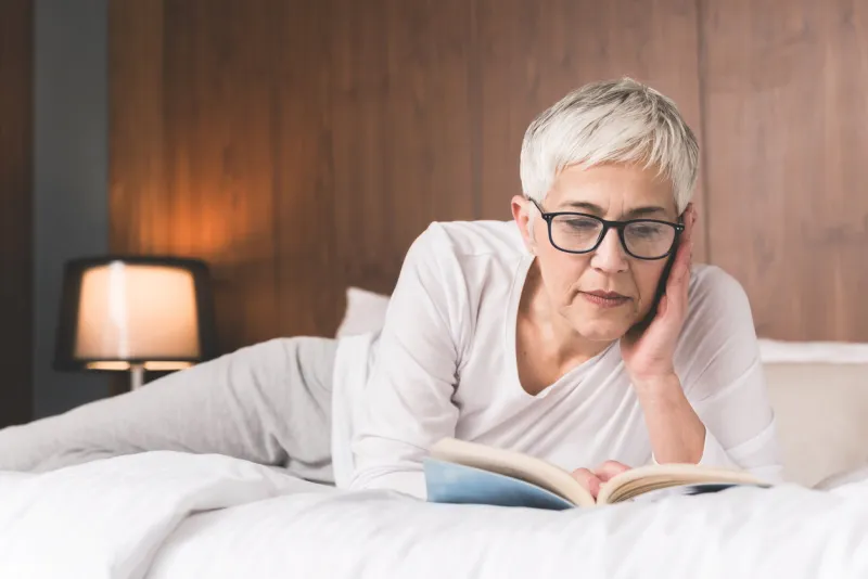 beautiful concentrated mature woman with glasses lying on a comfy bed and reading a book in her bright modern bedroom before sleep, literature and education concept