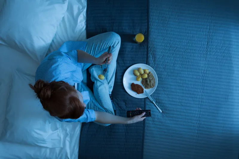 photo from the top of young girl sitting on a bed, eating dinner and watching television