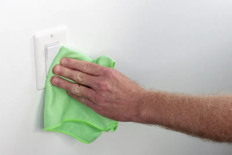 persons hand wiping a wall white light control inside white wall light switch cleaned with a cloth indoor flat white light switch being cleaned by a hand with a rag as part of house cleaning chores