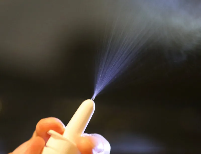 person's fingers pressing on a white nasal spray pump with mist being sprayed from the nozzle
