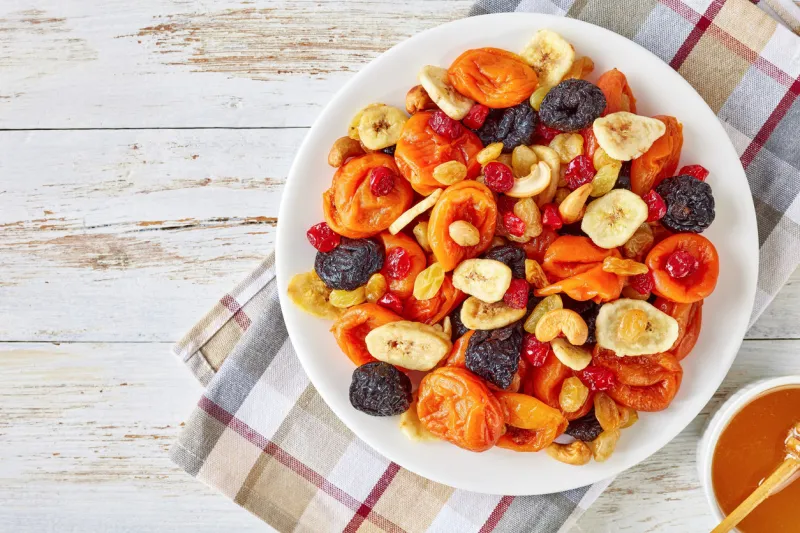 dried fruits and nut mix bowl - banana slices, apricots, raisins, prunes, cherries and cashew on a rustic table with honey in a bowl, horizontal view from above, close-up, flatlay