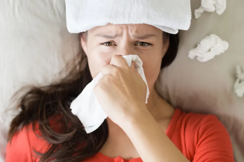 a woman resting on the bed with a wet towel on her head and tissue everywhere from blowing her nose flu season