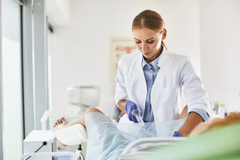 waist up portrait of gynecologist in white lab coat and sterile gloves using vaginal speculum during pelvic exam she is looking at instrument and smiling