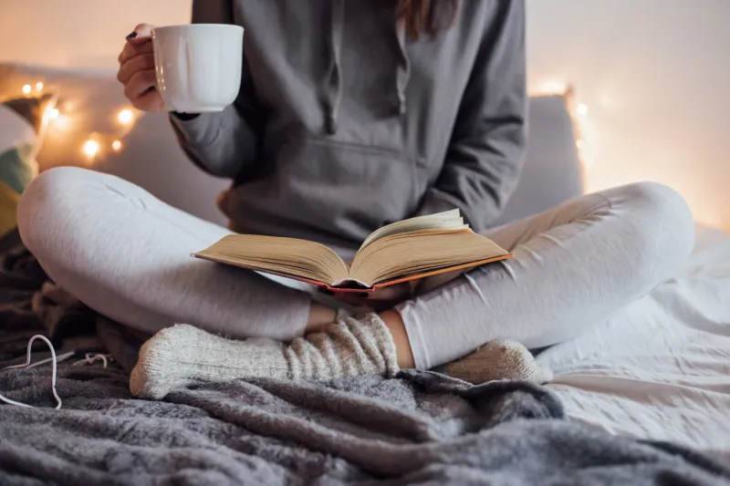 girl holding cup of hot tea and reading in bed around her in bad earphones, book, smart phone decorative lights in background