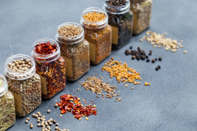 assortments of spices, white pepper, chili flakes, lemongrass, coriander and cumin seeds in jars on grey stone background copy space