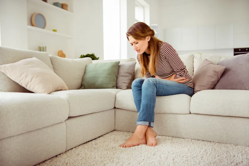 portrait of upset disappointed lady sit divan touch tummy had indigestion food poisoning hurt injury suffering dressed in denim outfit striped shirts close eyes long curly wavy hair in apartment