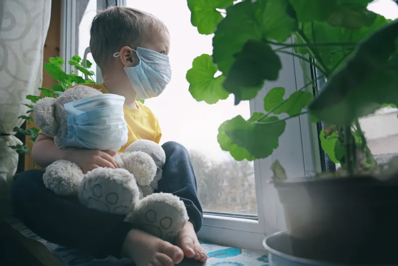 quarantine, threat of coronavirus, virus protection, pandemic child and his teddy bear both in protective masks sits on windowsill inside house and looks out the window